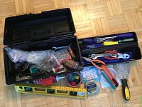 Black plastic toolbox opened showing trays with assorted hand tools including screwdrivers, pliers, tapes, level, and adhesive.