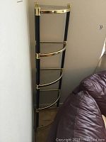 Full view of corner book stand with brass and black metal frame, four glass shelves, placed at corner of room next to bed.