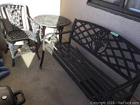 Photo showing black metal bench with lattice and floral design on backrest next to metal chair and small round table on porch.