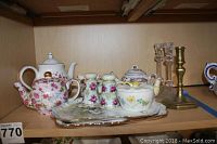 Porcelain teapot with pink rose floral pattern alongside creamer, sugar bowl, salt and pepper shakers on a tray, plus two glass and one brass candlestick holders on a wooden shelf.