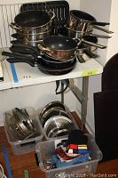 View of assorted pots and pans stacked on table including non-stick frying pans, stainless steel saucepans, and metal trays in plastic bins below.