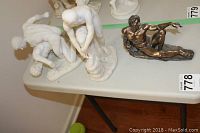 Three statues on a table; two white resin male nude statues and one bronze-tone male nude statue.
