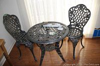 Photo of cast aluminum patio table and two chairs with floral and lattice design, positioned indoors on hardwood floor near windows.