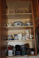 View of cupboard interior showing multiple shelves with assorted ceramic mugs, glass cups, decorative bowls and teapots.