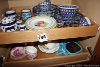 Assortment of decorative dishware in cabinet drawers including Boleslawiec blue hand-painted ceramics, pink and white tea cups, and assorted plates.