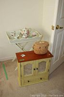 Photo showing wood wall cabinet with mirror on door, painted green with cherry stained top, placed near door. On and near the cabinet sit a woven basket and a white folding tea tray with floral tea set.