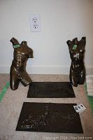 Photo showing two bronze-colored metal torso sculptures, male and female, positioned on carpeted floor against wall outlet.