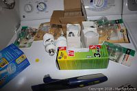 Light bulbs assortment including compact fluorescent and packaged bulbs with a green and yellow 3-way bulb box in front and a blue LED bulb pack to the side. Also includes a small electronic device that resembles a pen.