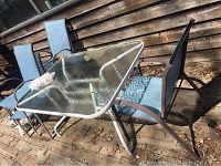 Glass top outdoor table with four blue mesh chairs placed on wooden deck against wooden fence.