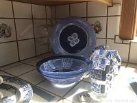 Two blue serving bowls stacked, blue serving plate behind them, and a group of 12 tea cups stacked in pairs and singles next to them. Items on a tiled kitchen surface.