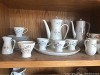 Two sets of teapots and tea cups displayed on wooden shelves, one set with blue/purple floral motif, the other with cream/pink/green floral motif, totaling 27 pieces.