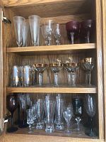 Multiple shelves of wood cabinet with various styles of wine glasses and glassware, including clear, colored, and gold-trimmed glasses.