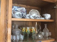 Wooden shelf holding Gem China tea cups, sugar bowl, creamer with black floral design on upper shelf. Below, clear glass decanters, various clear wine glasses, including hand-painted sunflower glasses.