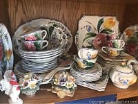 Overview photo showing multiple ceramic pieces with floral patterns, including cups, plates, bowls, and serving dishes arranged on a wooden shelf.