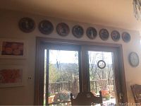 Wide view showing eight Norman Rockwell collectible plates mounted above sliding door, wall clock to right, and a small bead wall hanging on the wall.