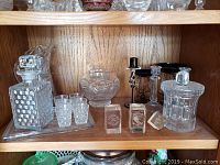 Shelf display of various crystal and glassware including decanter, glasses, bowl, paperweights, and candle holders.