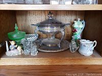 Shelf with various household decor including ceramic figurines, glass pieces, and silver-tone serving dish