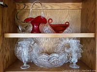 Wood cabinet shelf displaying clear patterned crystal bowl and two vases, red glass decorative pieces including a basket, pedestal vase, and oval bowl.