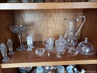 Shelf with assorted 29 pieces of crystal home decor including shakers, compote dish, small figurines, covered candy dish, pitcher, and dishes.