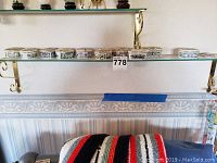 Photo of 7 white ceramic music boxes with various floral and animal paintings on a glass shelf with brass supports.