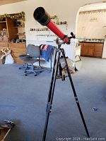 Red refractor telescope on black metal tripod inside a home.