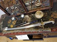 The full lot displayed on a wooden floral-decorated table with a mirrored ornate metal frame background. Includes the sword, sheath, knife, candle holders, and decorative jar.