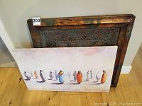 Metal artwork with a brass look and acrylic painting on canvas leaning against a wall, hardwood floor visible