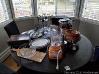 Full view of kitchen items on dark round table including KitchenAid blender, crystal fruit bowl, ceramic pot, pressure cooker, muffin pan, plates, cutting board, and racks.