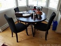 Full view of round wood dining table with four black slipcovered chairs around it, showing table and chair design.