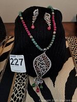 View of necklace with multicolor beads and intricate teardrop pendant along with matching earrings displayed on black stand.