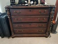 Front view of four drawer wood dresser with brass handles, resting on carpeted floor next to black suitcase.