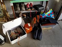 Overall lot showing multiple men's shoes and boots of various styles, a colorful shopping bag, black graduation gown, storage containers, cigar boxes, and a hair clipper.