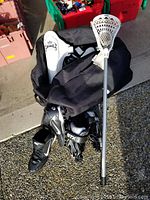 Photo of lacrosse stick, black bag, shoulder pads, and cleats arranged on ground