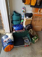 Camping gear including Coleman green two-burner stove, rolled sleeping bags in various colors, backpacks, and boxed tent in storage bags.