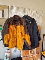 Photo showing a men's large yellow and brown The North Face jacket next to a dark women's jacket against a pegboard.