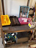 Gray metal shelf with plastic organizers containing assorted hardware, screws, and small tools. Bottom shelf holds a black hard case and various other tools.