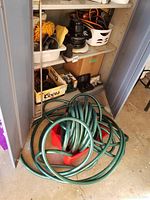 Storage cabinet shelf with black and cream orbital car waxer and attachments. Nearby car care items in boxes and baskets on shelf. Green garden hose coiled on red holder on floor.