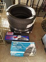 Two Pirelli tires stacked with Quality brand tire chains in a black carrying case and Laclede truck cable chains box placed nearby on floor.