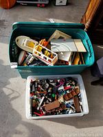 Two plastic bins on a floor, one containing a white and yellow Lego boat partially assembled alongside various other Lego pieces, and the other bin filled with assorted loose Lego bricks and parts, showing a large mix of colors including brown roof pieces.