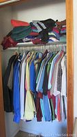 Closet with multiple men's button-down shirts hanging on metal rod, tops of shelf with folded sweatpants and clothing in various colors.