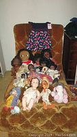 Photo showing wide assortment of vintage dolls on a brown upholstered chair including dark-skinned vinyl dolls, various small dolls, a large cloth doll, and a plush bunny.