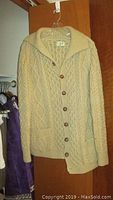 Men's beige Irish wool cable knit cardigan with large buttons and shawl collar, hanging on wooden door.