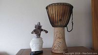 Djembe drum with carved body and dancing woman figurine shown side by side, both against plain wall