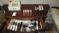 Photo of a wooden drawer with various stainless steel flatware organized inside and on top, including forks, knives, spoons, and collectible Jack Daniel's drink stir sticks.