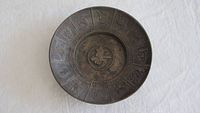 Top-down photo of a shallow brass bowl with engraved symbols around the rim and center character, dark patina visible.