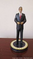 Photo of Barack Obama figurine showing full body, dark suit, red tie, standing on base with inscription