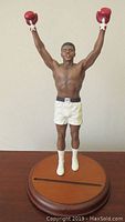 Front view of 11-inch Muhammad Ali sculpture in a boxing victory pose with arms raised, wearing white shorts, red gloves, and white boots on wooden base.
