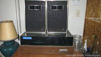 Photo of two vintage black speakers placed on top of the turntable wood casing along with a green lamp and some glassware adjacent.