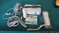 Photo showing the Nintendo Wii console components on a green surface including the console, power adapter, sensor bar, composite AV cable, Wii Remote controller with wrist strap, Wii Sports game, and a small dog toy.