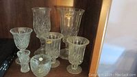 Group of five clear lead crystal items including vases and candle holders, positioned on a wooden shelf. Items vary in size and design.